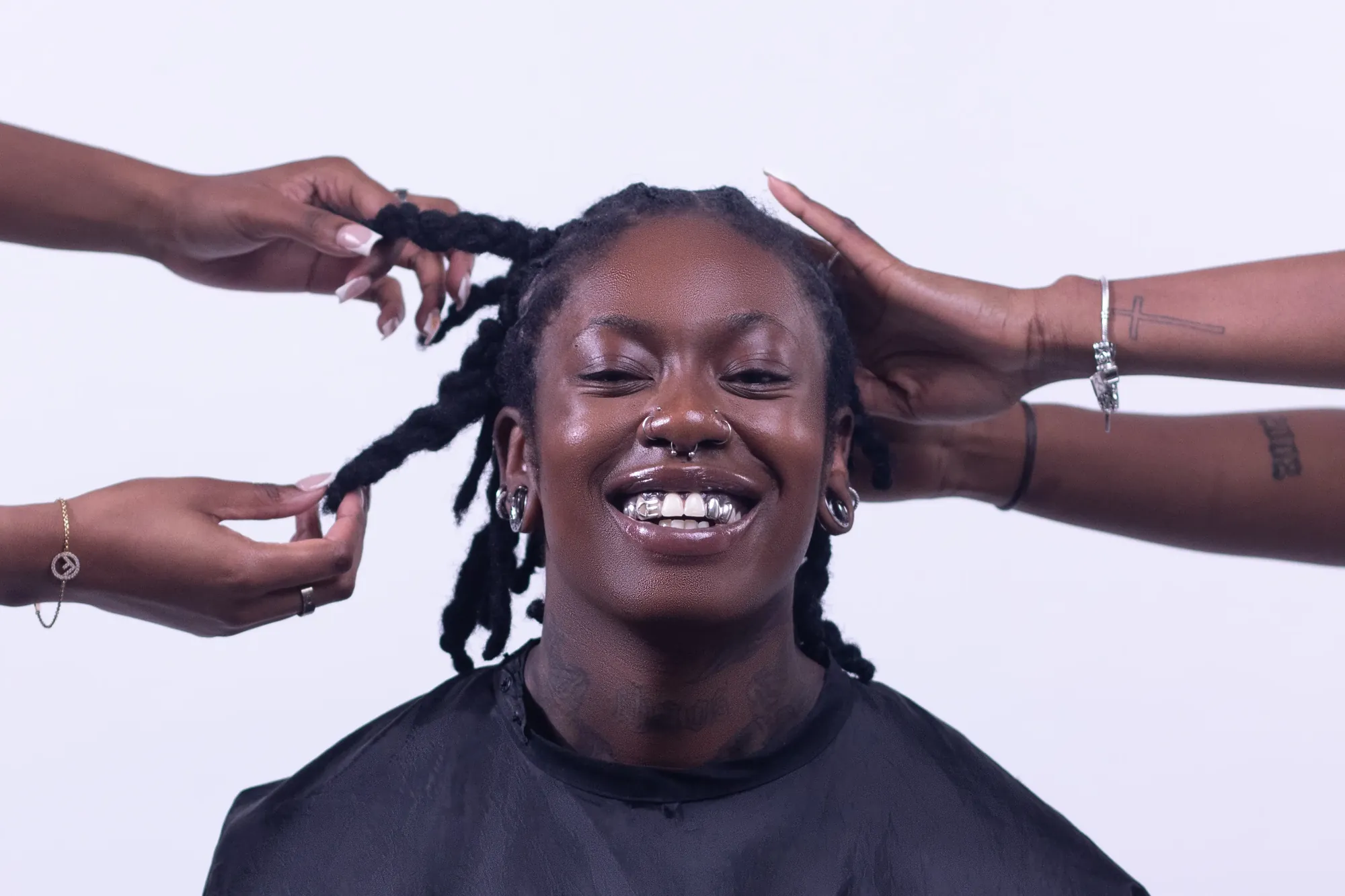 Several hands styling a smiling client’s locs